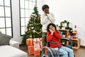 Young interracial couple with woman sitting on wheelchair by christmas tree sleeping tired dreaming and posing with hands together