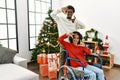 Young interracial couple with woman sitting on wheelchair by christmas tree smiling making frame with hands and fingers with happy Royalty Free Stock Photo