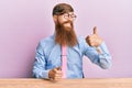 Young irish redhead man wearing business shirt and tie sitting on the table looking proud, smiling doing thumbs up gesture to the Royalty Free Stock Photo