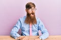 Young irish redhead man wearing business shirt and tie sitting on the table in shock face, looking skeptical and sarcastic, Royalty Free Stock Photo