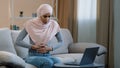 Young islamic arab business woman muslim girl in hijab sitting on sofa using laptop at home working studying online Royalty Free Stock Photo