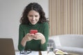 Young italian freelancer lady with dark wavy hair holding cell phone and looking at screen with smile