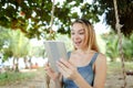 Young jocund woman using tablet and riding swing on sand, wearing jeans sundress. Royalty Free Stock Photo