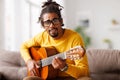 Young joyful african american man playing acoustic guitar at home, sitting on sofa in living room Royalty Free Stock Photo