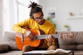 Young joyful african american man playing acoustic guitar at home, sitting on sofa in living room