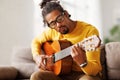 Young joyful african american man playing acoustic guitar at home, sitting on sofa in living room