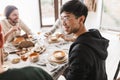 Young joyful asian man with dark hair in eyeglasses and hoodie sitting at the table happily looking aside. Group of Royalty Free Stock Photo