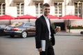 Young joyful businessman in black suit and white shirt with wireless earphones holding laptop and coffe in hands while