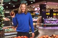 Young joyful guy in dark blue sweater happily looking in camera while holding mango in hand in modern supermarket