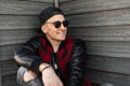 Young joyful hipster man with a cute smile in a stylish checkered jacket in a trendy cap in stylish sunglasses is sitting