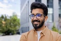 Young joyful man close-up smiling and looking away, Indian man in glasses outside office building walking in the city Royalty Free Stock Photo