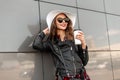 Young joyful woman with red lips and sweet smile in hat in black sunglasses and leather jacket with tasty latte posing and smiles