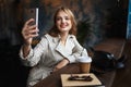 Young joyful woman in striped trench coat happily taking photos on cellphone with little black handbag and coffee to go Royalty Free Stock Photo