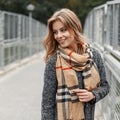 Young joyful woman with a sweet smile in a trendy coat with a trendy checkered scarf walks down the street