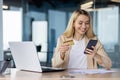 Young joyful woman at workplace inside office holds phone , bank credit debit card in hands, businesswoman chooses gifts