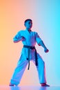 Young karateka in a defensive posture, teen boy practicing against gradient orange blue background in neon light