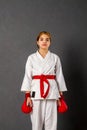 Young karateka girl in a white kimono and red competition outfit trains and performs a set of exercises against a gray wall Royalty Free Stock Photo