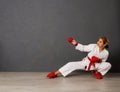 Young karateka girl in a white kimono and red competition outfit trains and performs a set of exercises against a gray wall Royalty Free Stock Photo