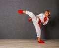 Young karateka girl in a white kimono and red competition outfit trains and performs a set of exercises against a gray wall Royalty Free Stock Photo