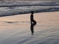 A young kneeling boy daydream and enjoying alone the wet beach sand at sunset time