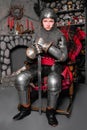 A young knight in medieval armor with a sword in his hand sits in a rocking chair by the fireplace