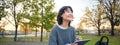 Young korean woman sits in park, draws on digital tablet, writes in gadget with pen tool, looks around and makes notes