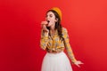 Young lady in fluffy white skirt bites donut. Parisian in beret and yellow blouse posing on red background. Royalty Free Stock Photo