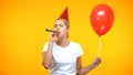 Young lady in party hat blowing horn, holding red balloon, birthday celebration