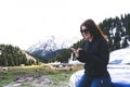 young lady sitting on a bonnet of a car using smart phone in the mountains. Happy Girl sending message with sun glasses Royalty Free Stock Photo