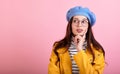 A young lady in a stylish hat and beret and a yellow raincoat with glasses shows thinking on a pink background Royalty Free Stock Photo