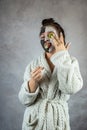 Young lady in white bathrobe applying white and black clay masks on face and holding cucumber slice Royalty Free Stock Photo