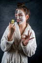 Young lady in white bathrobe applying white and black clay masks on face and holding cucumber slice Royalty Free Stock Photo