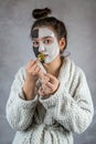 Young lady in white bathrobe applying white and black clay masks on face and holding cucumber slice Royalty Free Stock Photo