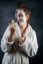 Young lady in white bathrobe applying white and black clay masks on face and holding cucumber slice Royalty Free Stock Photo