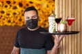 Young Latin American waiter serving cocktails on a tray. He wears a medical mask