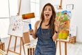 Young latin artist woman holding paint roller standing at art studio celebrating victory with happy smile and winner expression Royalty Free Stock Photo