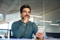 Young Latin business man in office holding smartphone looking away thinking. Royalty Free Stock Photo