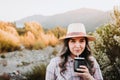 Smiling woman with a hat and wearing a pale pink blouse, drinking mate in a natural space at sunset