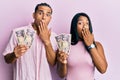 Young latin couple holding japanese yen banknotes covering mouth with hand, shocked and afraid for mistake