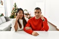 Young latin couple sitting on the table by christmas tree mouth and lips shut as zip with fingers Royalty Free Stock Photo