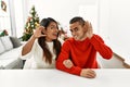 Young latin couple sitting on the table by christmas tree smiling with hand over ear listening an hearing to rumor or gossip
