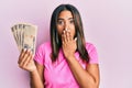 Young latin girl holding japanese yen banknotes covering mouth with hand, shocked and afraid for mistake