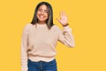 Young latin girl wearing wool winter sweater waiving saying hello happy and smiling, friendly welcome gesture