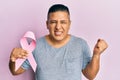 Young latin man holding pink cancer ribbon screaming proud, celebrating victory and success very excited with raised arm Royalty Free Stock Photo