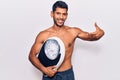 Young latin man shirtless holding weighing machine pointing finger to one self smiling happy and proud