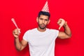 Young latin man wearing birthday hat with angry face, negative sign showing dislike with thumbs down, rejection concept