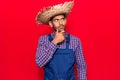 Young latin man wearing farmer hat and apron with hand on chin thinking about question, pensive expression