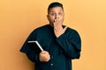 Young latin priest man holding bible covering mouth with hand, shocked and afraid for mistake