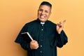 Young latin priest man holding bible smiling happy pointing with hand and finger to the side