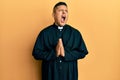 Young latin priest man praying with hands together angry and mad screaming frustrated and furious, shouting with anger looking up Royalty Free Stock Photo
